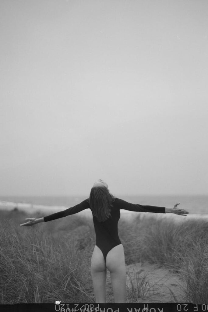 Woman in bodysuit embraces freedom with arms outstretched on a grassy beach.
